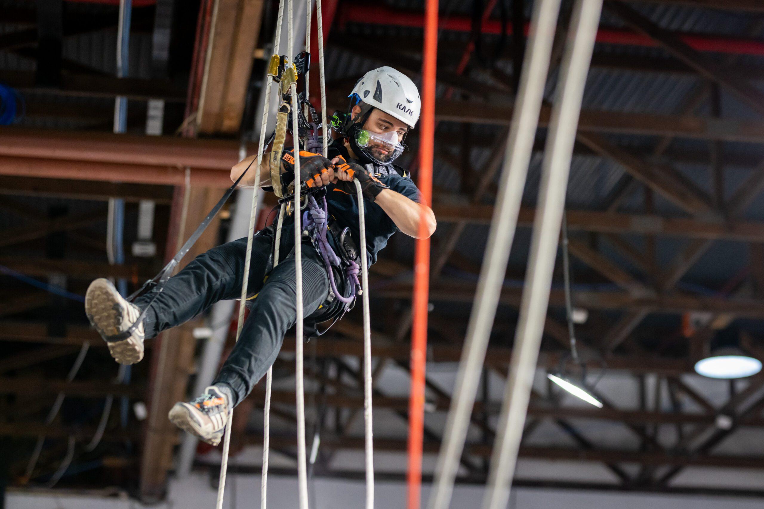 Helmeted rope-access technician wearing eye protection and a harness while working inside a building